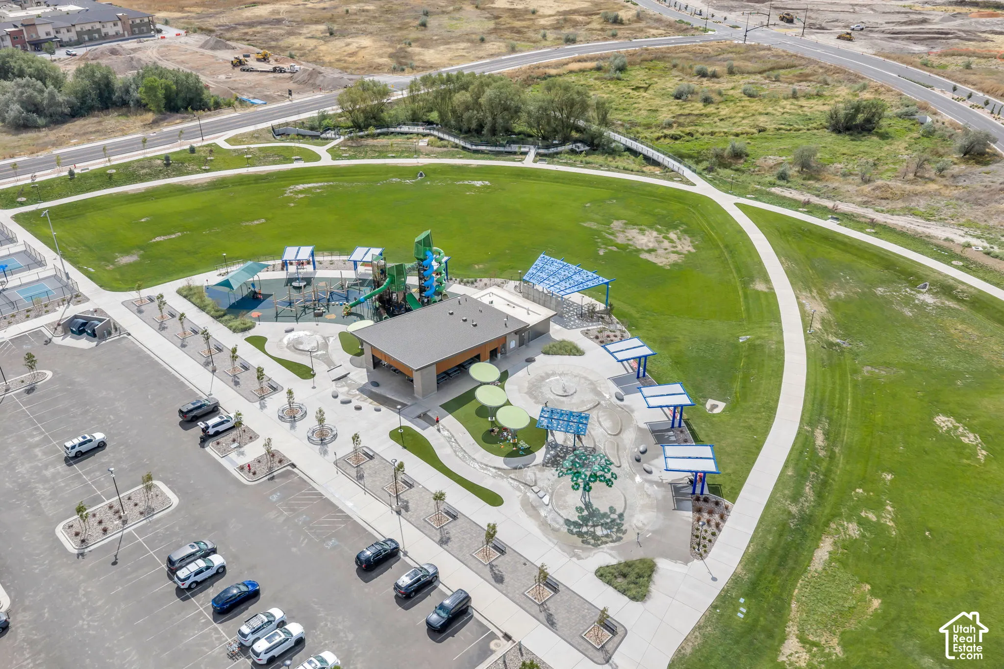 Aerial view of community recreational space featuring a paved parking lot, a central clubhouse structure, multiple covered seating areas, concrete walking paths, and expansive landscaped lawn