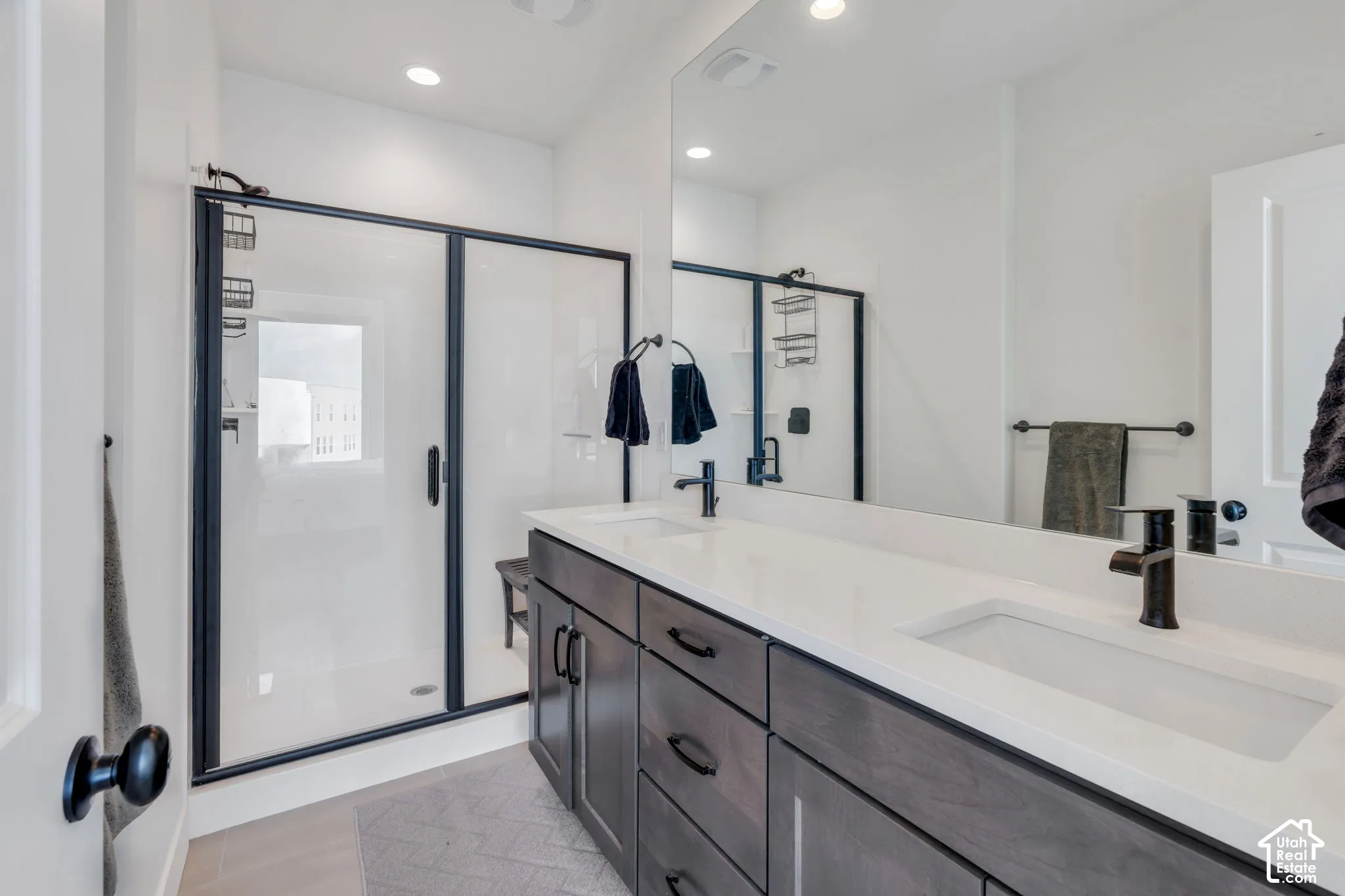 Double-sink vanity with matte black fixtures, quartz countertop, dark cabinetry, and a walk-in shower with a glass enclosure