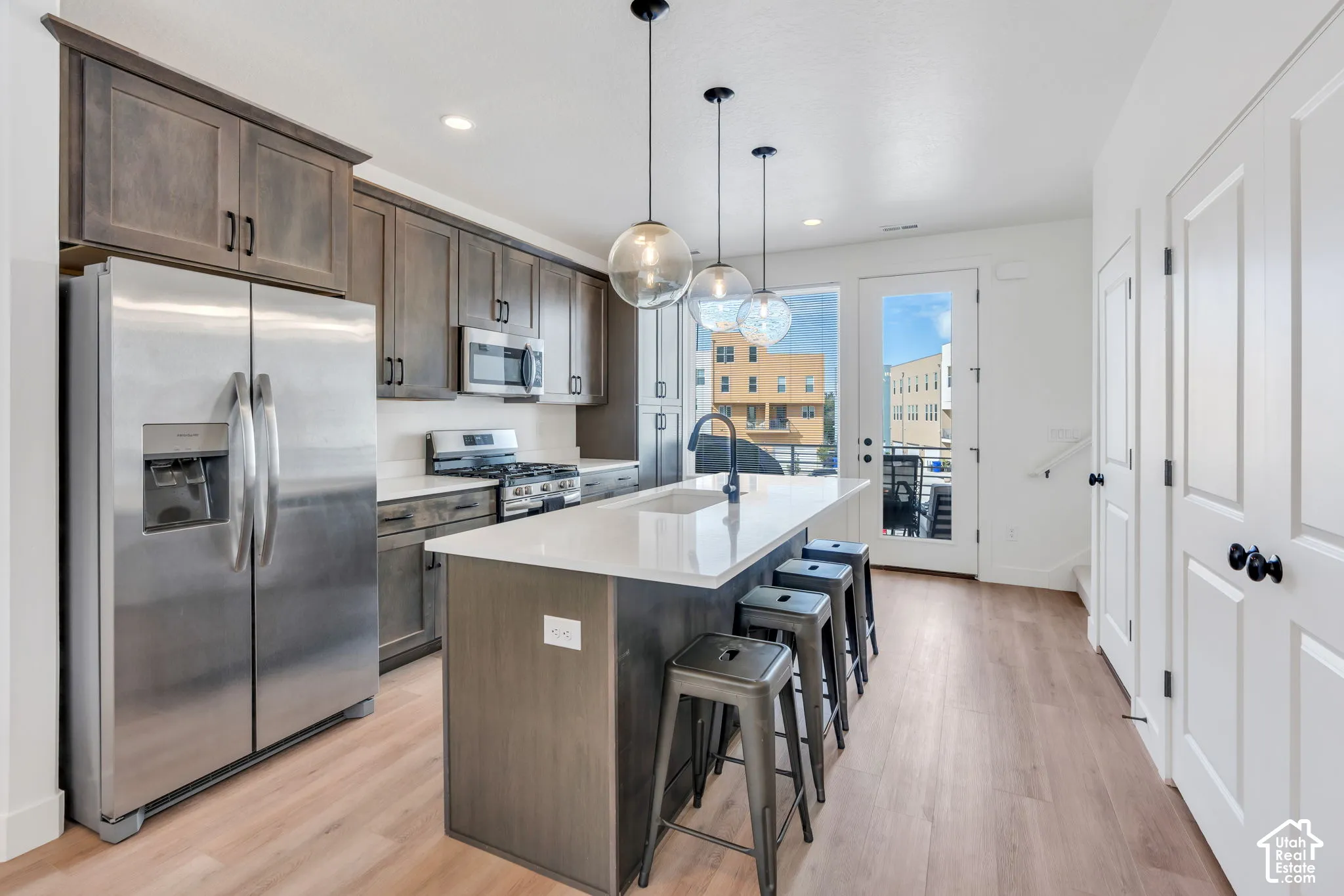 Kitchen featuring dark-toned cabinetry, stainless steel appliances, quartz countertops, a central island with a built-in sink, and wood-finish flooring