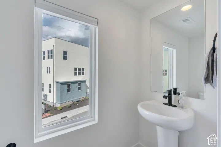 White pedestal sink featuring a matte black faucet, large frameless wall mirror, and window with adjustable roller shade