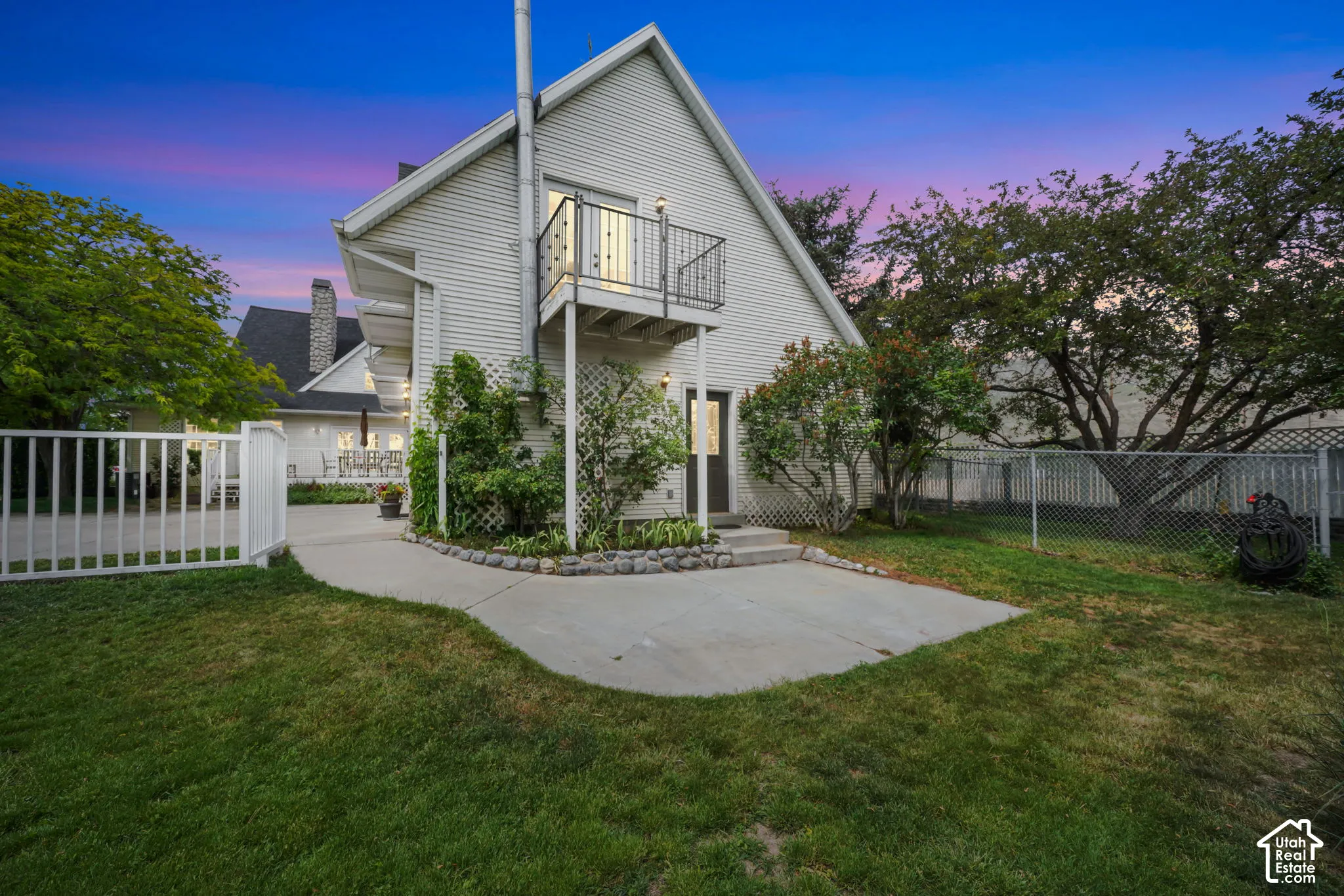 Exterior featuring white horizontal siding, a second-story balcony with metal railing, a concrete patio, and a chain-link perimeter fence