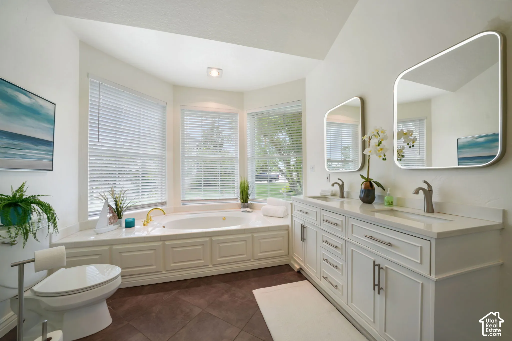 Double vanity with rectangular undermount sinks, stone countertops, and LED-backlit mirrors, complemented by a drop-in soaking tub set within a tiled surround, all featuring large bay windows and tile flooring