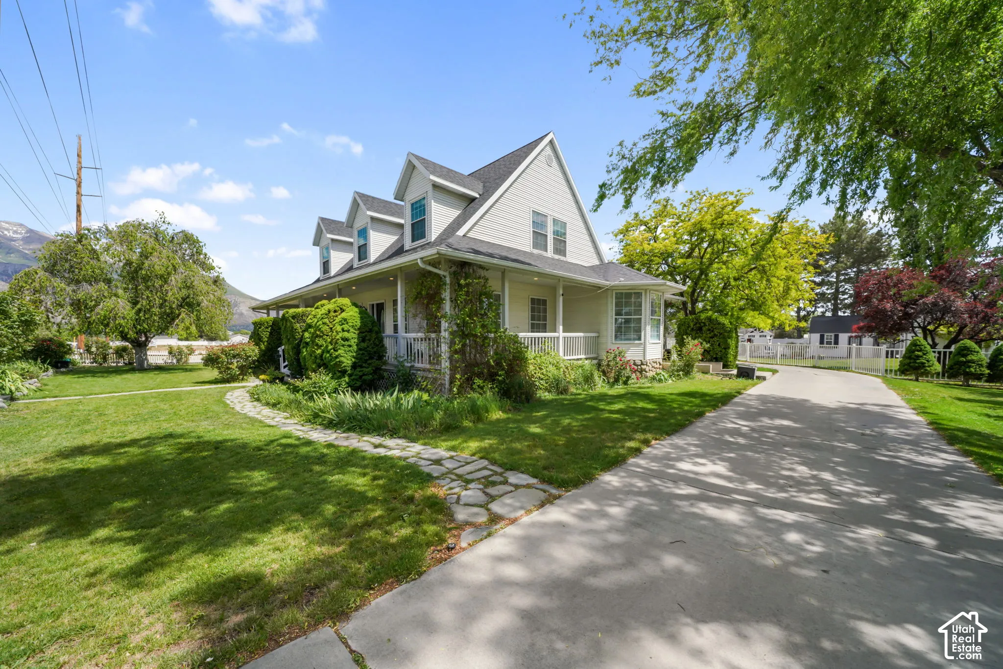 Two-story residence featuring light-toned horizontal siding, a wraparound covered porch with railing, multiple dormer windows, and a paved driveway leading to a stone walkway