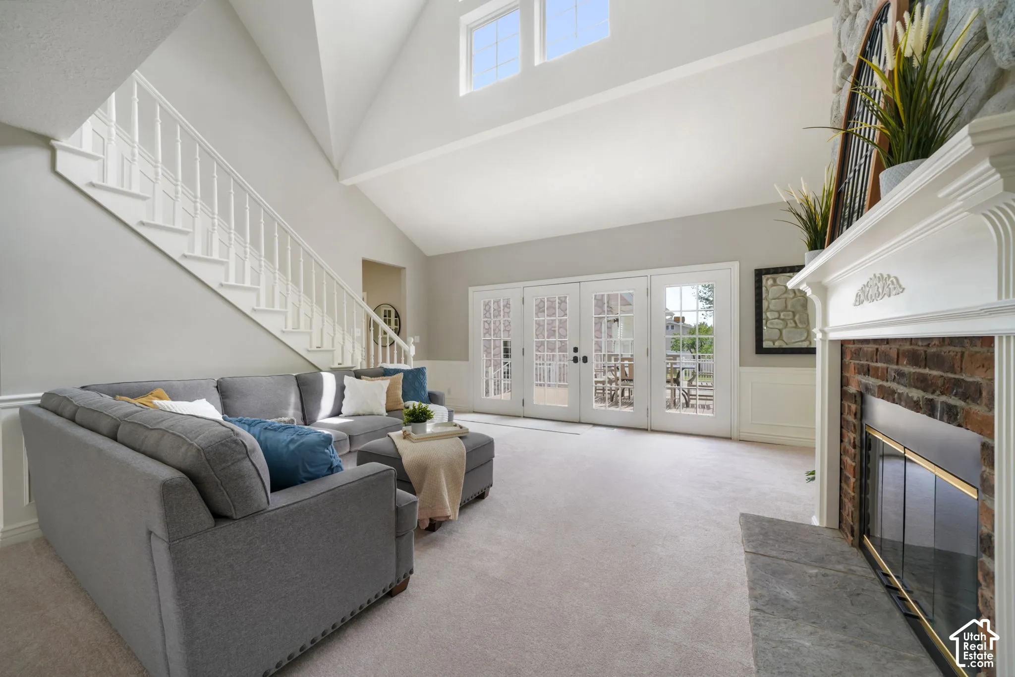 Vaulted ceilings, brick fireplace with decorative mantel, stone hearth, staircase with white railing, and glass-paneled double doors