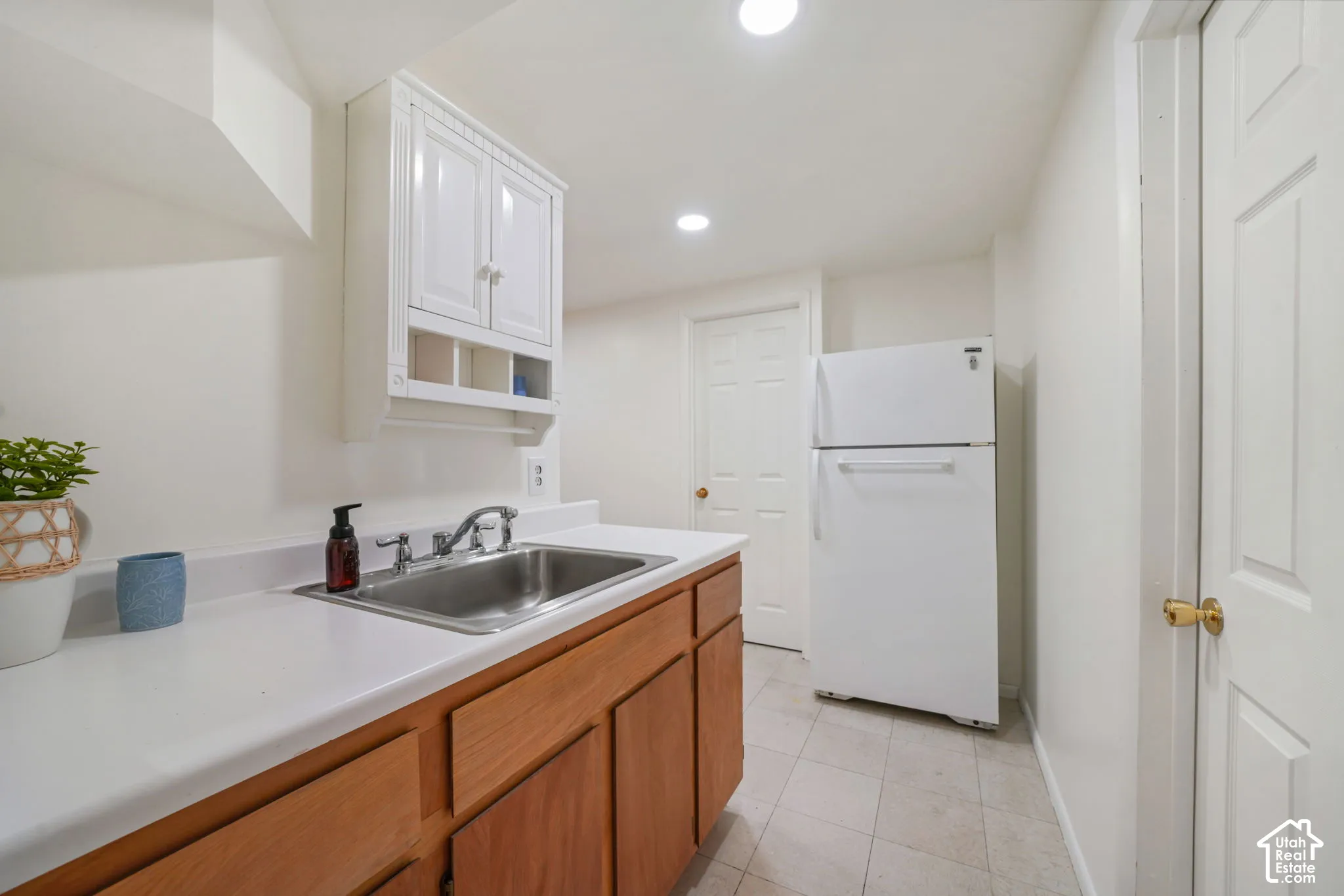 Stainless steel drop-in sink with chrome faucet, white countertop, wood-finish cabinetry, white wall-mounted storage cabinet, and neutral tile flooring