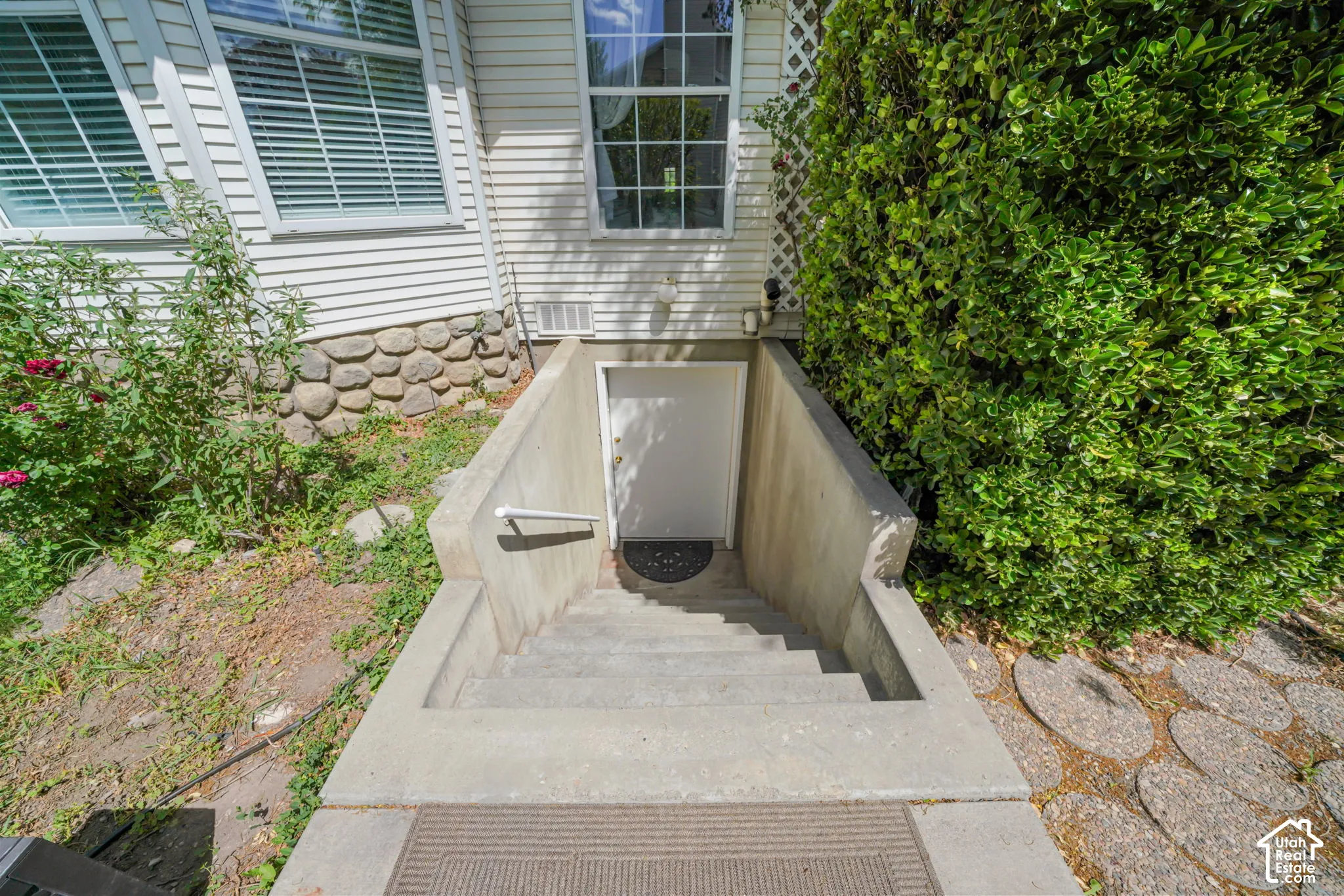 Exterior stairwell entrance with concrete retaining walls, white steel door, and metal handrail, finished with horizontal siding and stone foundation detail