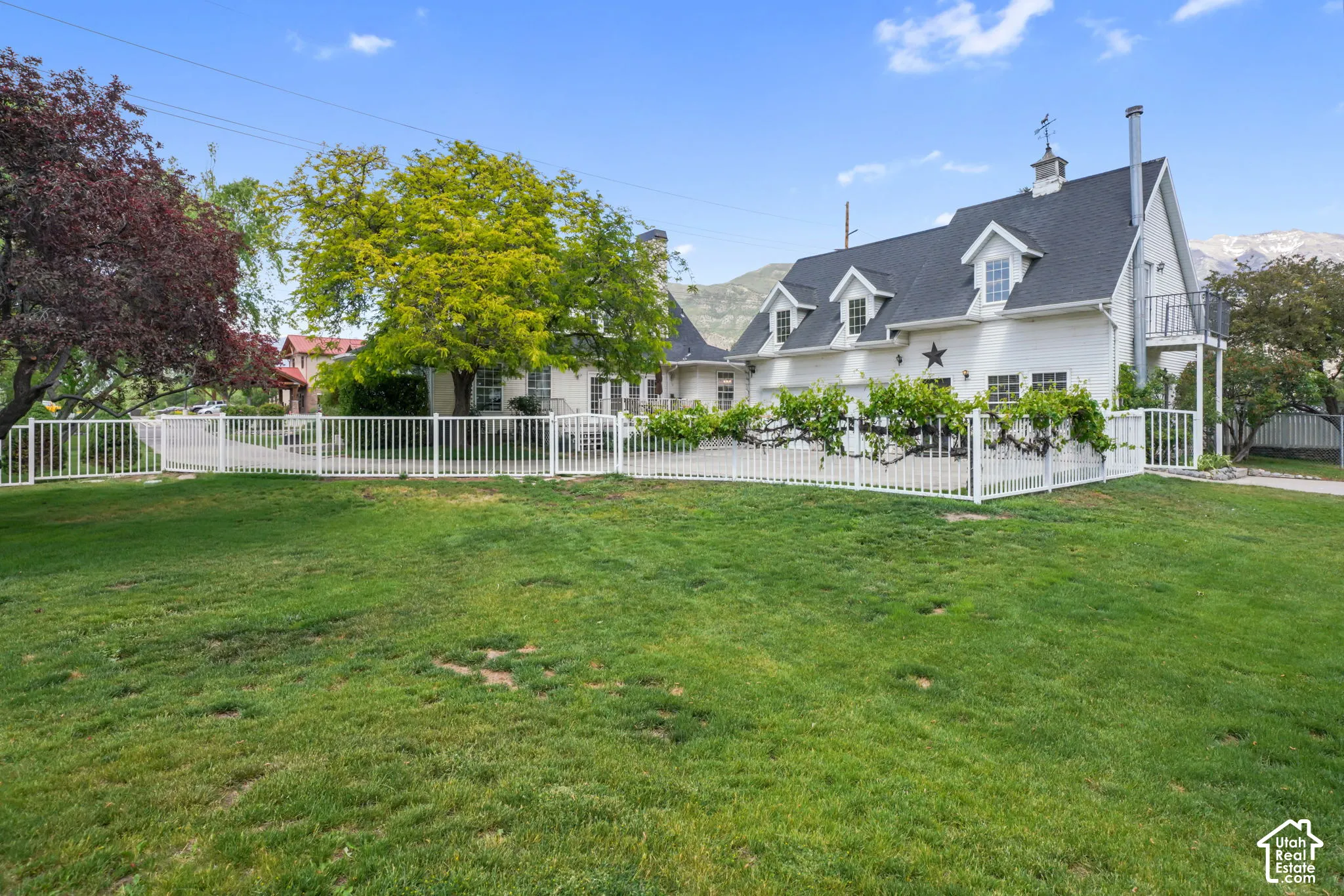 Exterior of the residence featuring white horizontal siding, steep-pitched roofline with dormers, attached second-story balcony, expansive grassy yard, and full-perimeter metal fencing