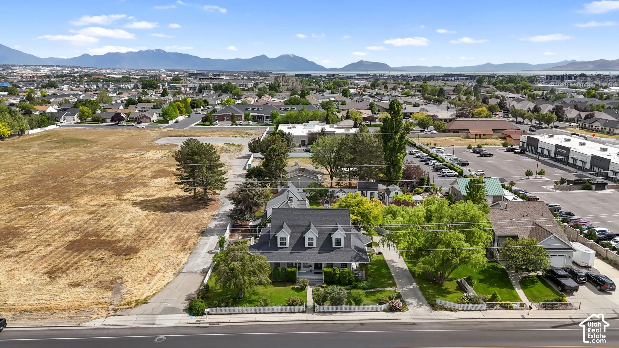 Aerial perspective featuring a multi-story residence with a pitched shingle roof, dormer windows, a covered front porch, and mature trees, set against a backdrop of mountain ranges