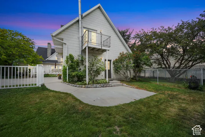 Exterior featuring white horizontal siding, a second-story balcony with metal railing, a concrete patio, and a chain-link perimeter fence