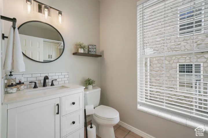 White vanity with stone-style countertop and undermount sink, subway tile backsplash, oval mirror with black frame, three-light vanity fixture, and horizontal blind window treatment