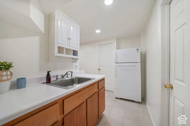 Stainless steel drop-in sink with chrome faucet, white countertop, wood-finish cabinetry, white wall-mounted storage cabinet, and neutral tile flooring