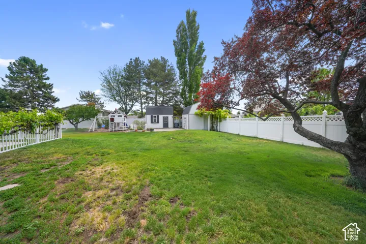 Spacious backyard featuring a manicured lawn, white vinyl fencing, mature trees, and detached storage structures