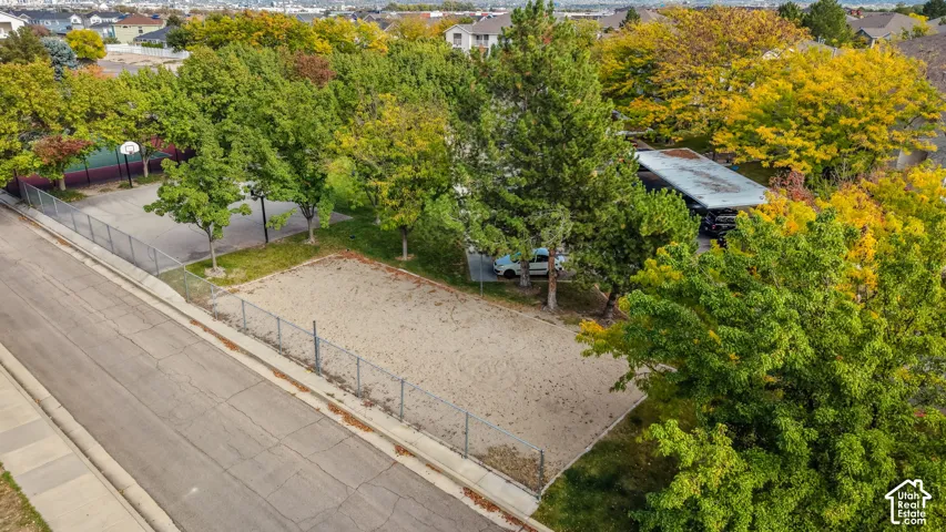 Outdoor recreational space featuring a fenced sand lot, paved court with a basketball hoop, covered parking structure, and mature trees