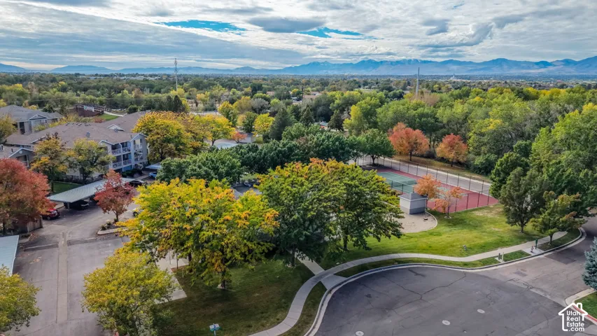 Aerial perspective of a multi-unit complex featuring paved parking areas with covered carports, a fenced tennis court, mature canopy trees, and mountain scenery