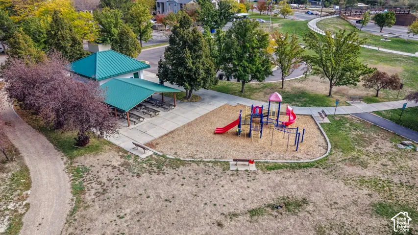 Community park area featuring a covered pavilion with metal roofing, concrete pathways, a structured playground set, and mature landscaping