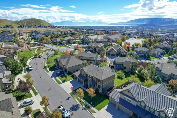 Aerial perspective featuring suburban residential development, multi-story dwellings, mature trees, manicured lawns, and prominent mountain and lake background scenery