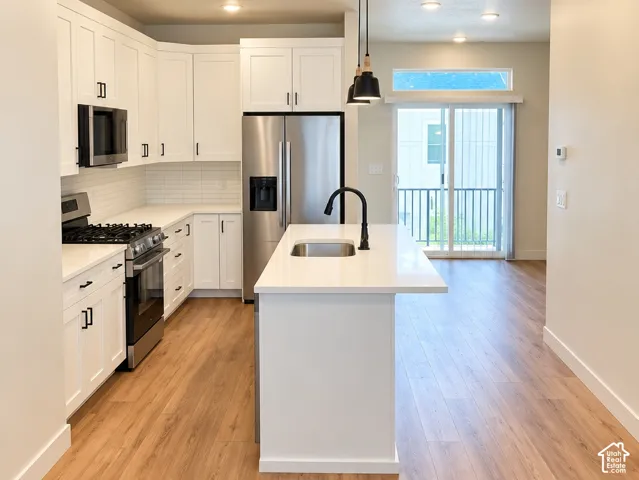 Kitchen featuring white shaker-style cabinetry, an island with a stainless steel undermount sink, quartz countertops, stainless steel appliances, and wood-finish flooring