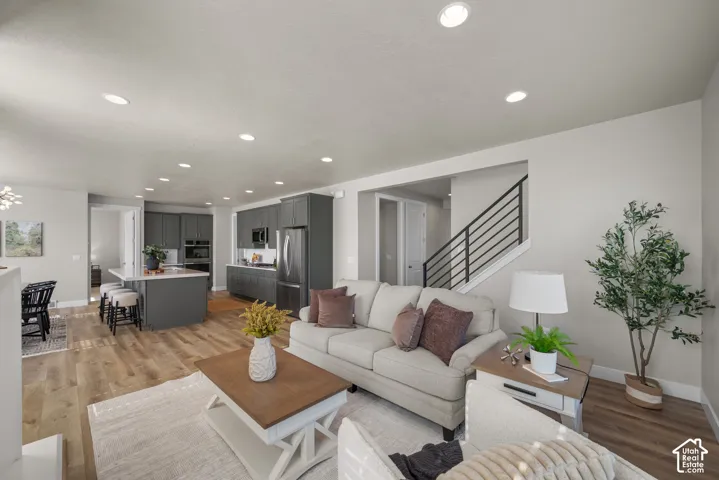 Open-concept living space featuring wood-finish flooring, recessed lighting, a kitchen island with stone-surface countertop, dark-toned cabinetry, and a staircase with black horizontal metal railings