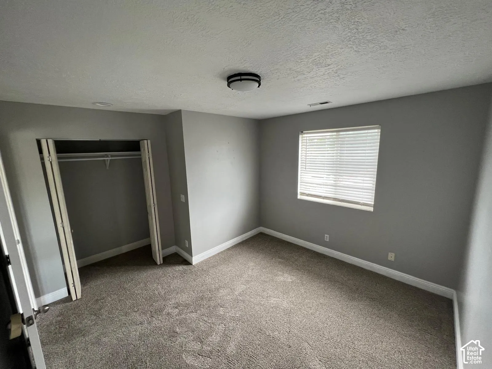 Neutral-toned interior featuring wall-to-wall carpeting, a recessed closet with bi-fold doors, a single-pane window with integrated blinds, and a flush-mount ceiling fixture