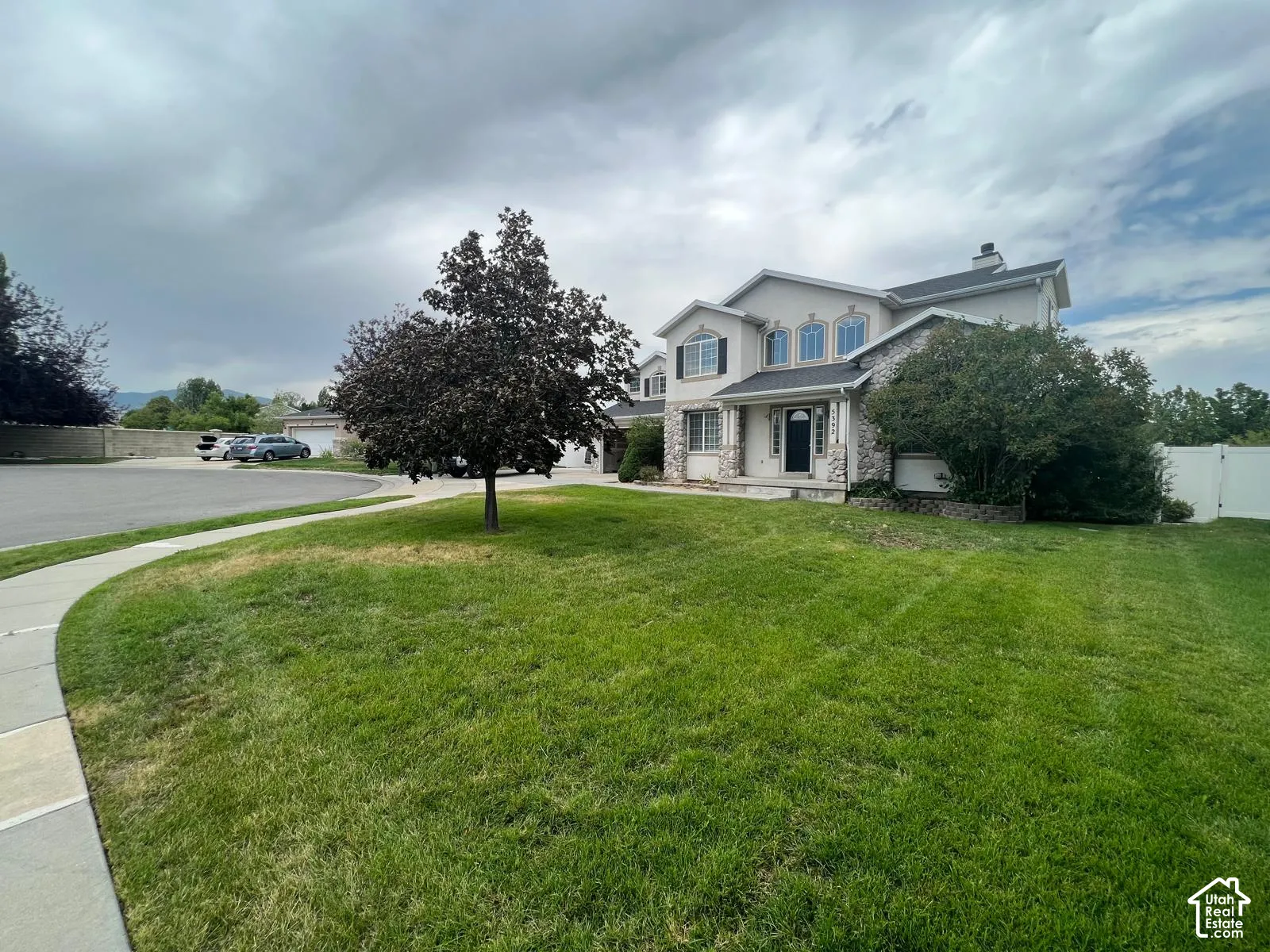 Two-story residence featuring light-toned exterior cladding, multi-textured stone veneer, arched window detailing, and a multi-gabled roofline, situated on a corner lot with an established lawn