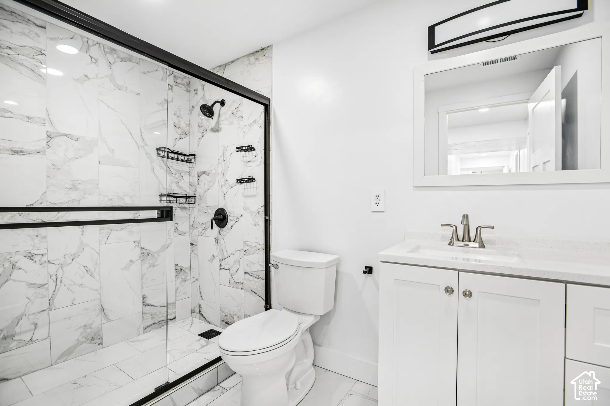 Bathroom featuring a walk-in shower with matte black hardware, stone-pattern tile surrounds, a white vanity with a composite countertop, and an integrated vanity mirror with task lighting