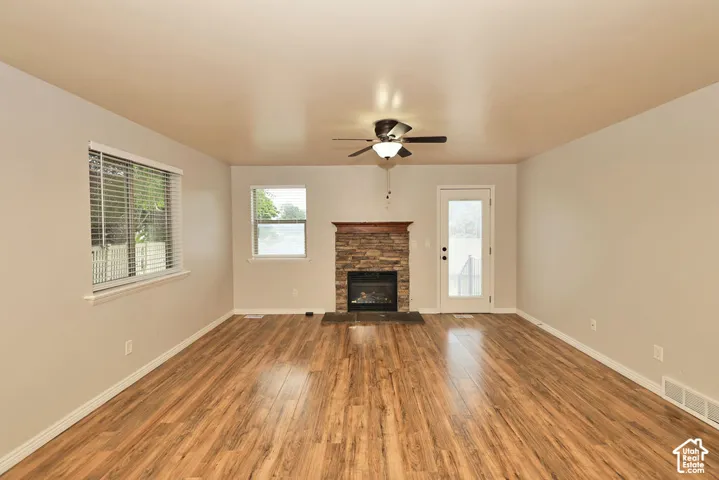 Living area featuring wood-finish flooring, stone-veneer fireplace with a wood mantel, ceiling fan with light fixture, and multiple windows