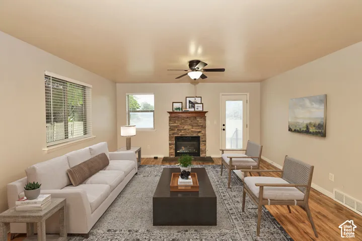 Stone-veneer fireplace with a wood mantel, wood-finish flooring, ceiling fan with light fixture, and large windows