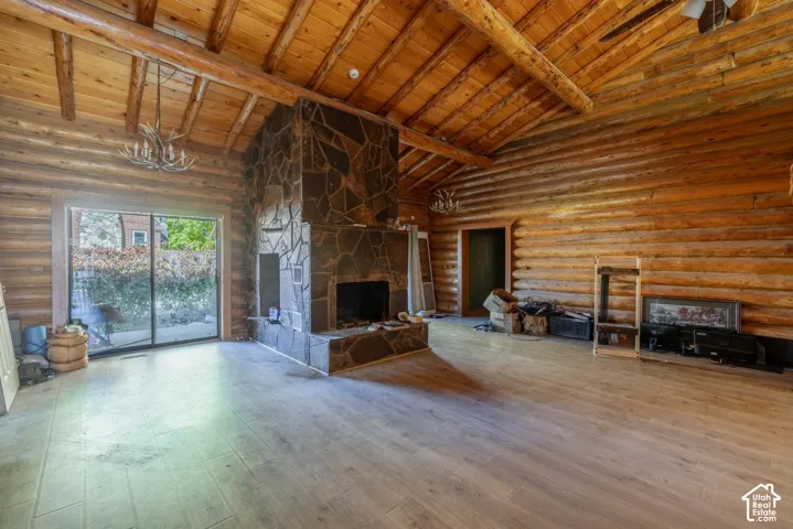 Vaulted timber ceiling with exposed log beams, full-log walls, large stone fireplace with hearth, wood-finish flooring, and sliding glass patio door