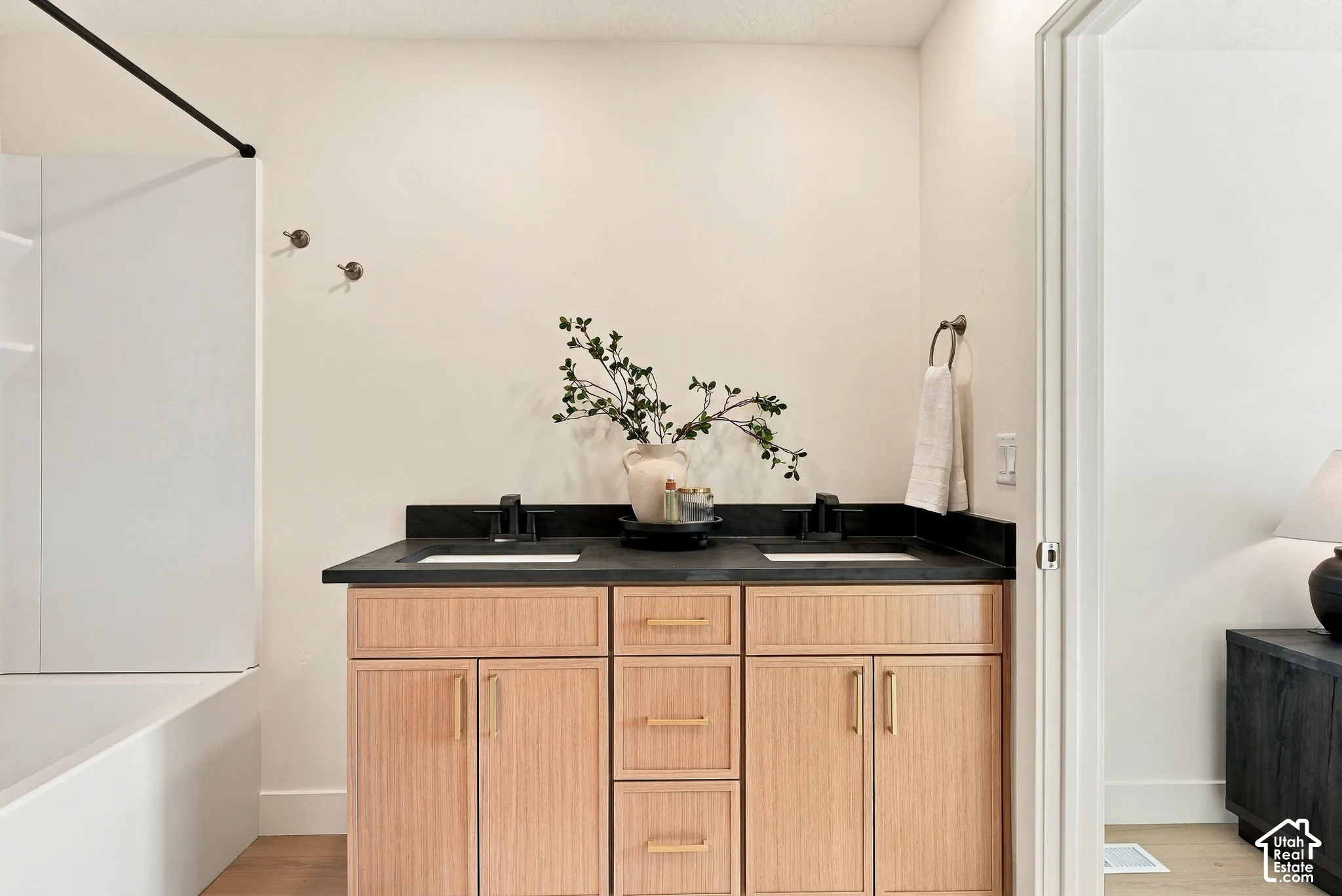 Double vanity featuring light wood-finish cabinetry, a dark stone-surface countertop, dual drop-in sinks, and matte black hardware
