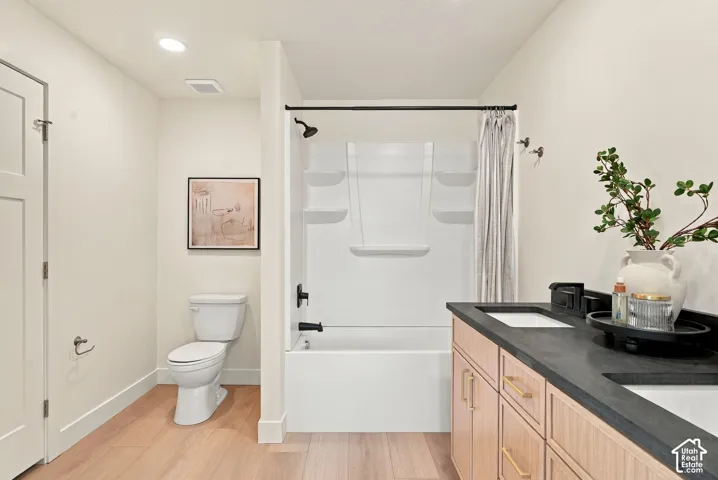 Bathroom featuring wood-finish flooring, dual-sink vanity with dark stone-style countertops, shaker-style cabinetry with gold-tone hardware, and a white tub-shower combination with matte black fixtures