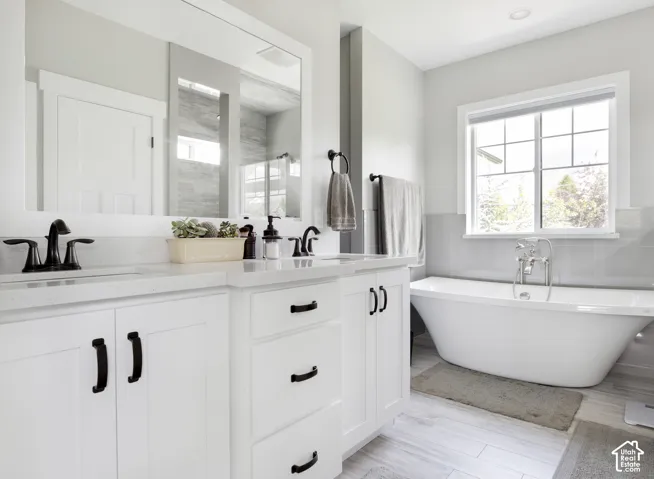 White shaker-style cabinetry with matte black hardware, dual undermount sinks, stone-finish countertops, freestanding soaking tub, and wood-finish flooring