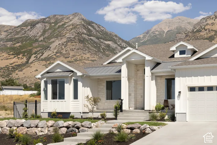 Exterior featuring white vertical siding, light-toned stone masonry, gabled rooflines with metal accents, and concrete stairs leading to a covered entry, set against a backdrop of mountainous terrain