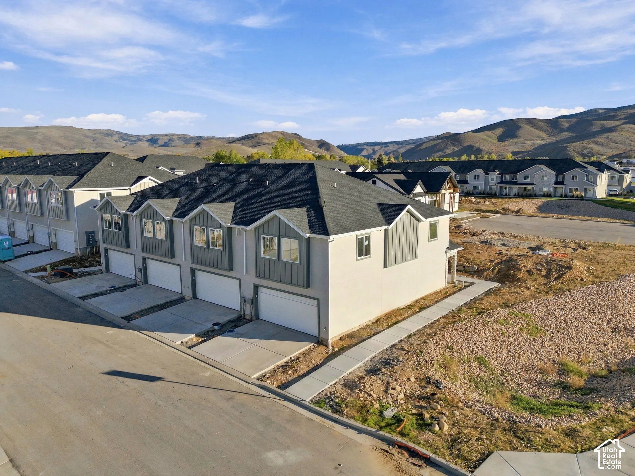 Multi-unit residential exterior featuring a light-colored facade, vertical paneling accents, asphalt shingle roofing, multi-car garages, and a mountainous backdrop