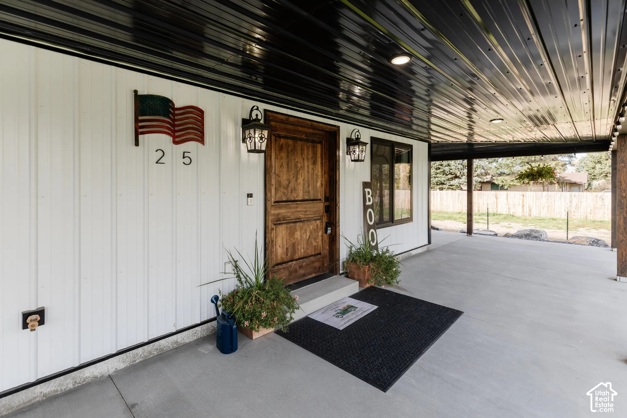 Covered patio featuring white vertical siding, black metal ceiling paneling, recessed lighting, and a stained wood entry door with wall-mounted lanterns