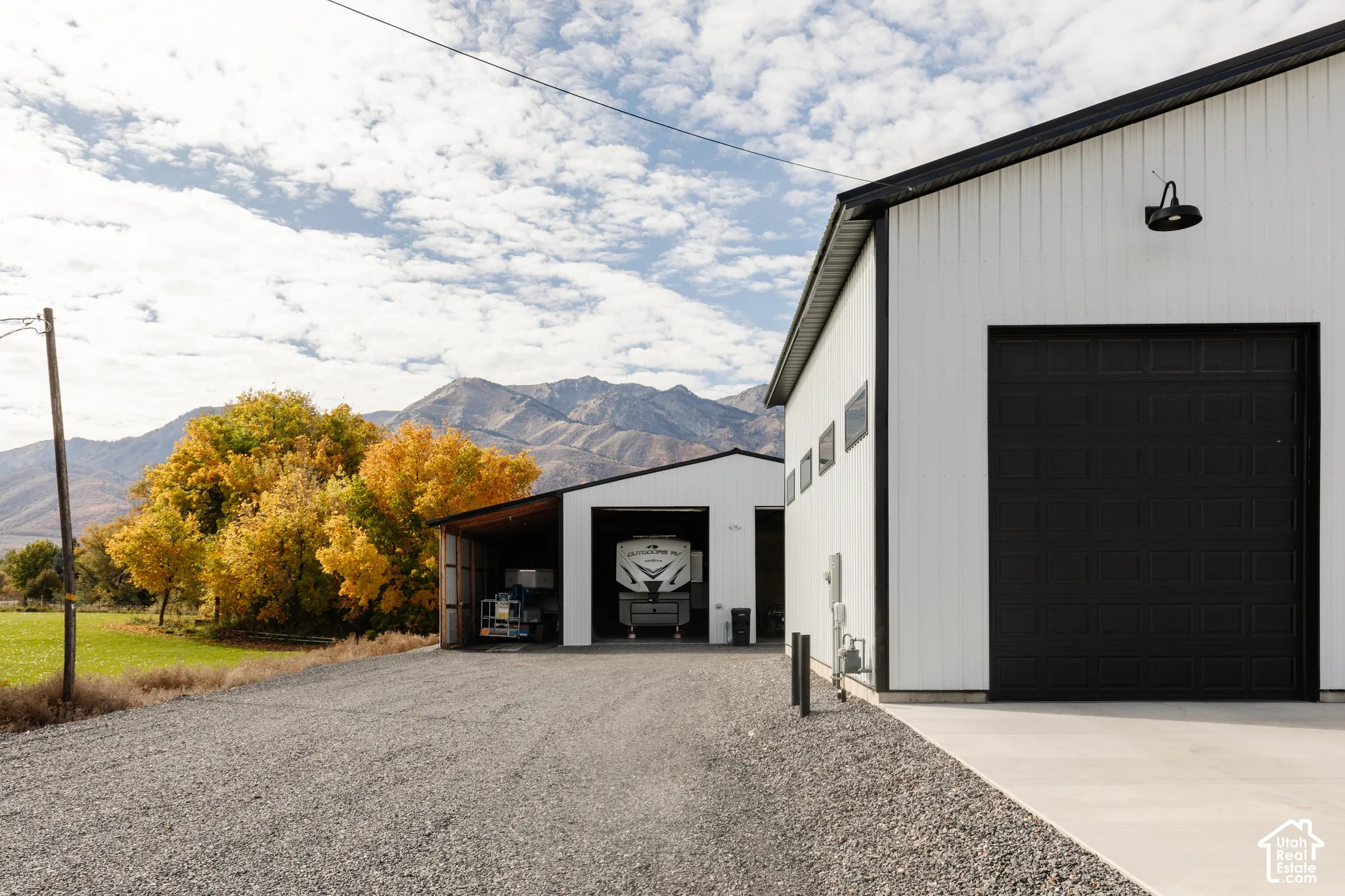 Metal-sided detached structures featuring high-clearance overhead garage doors, exterior wall-mounted gooseneck lighting, a gravel driveway, and a mountain backdrop