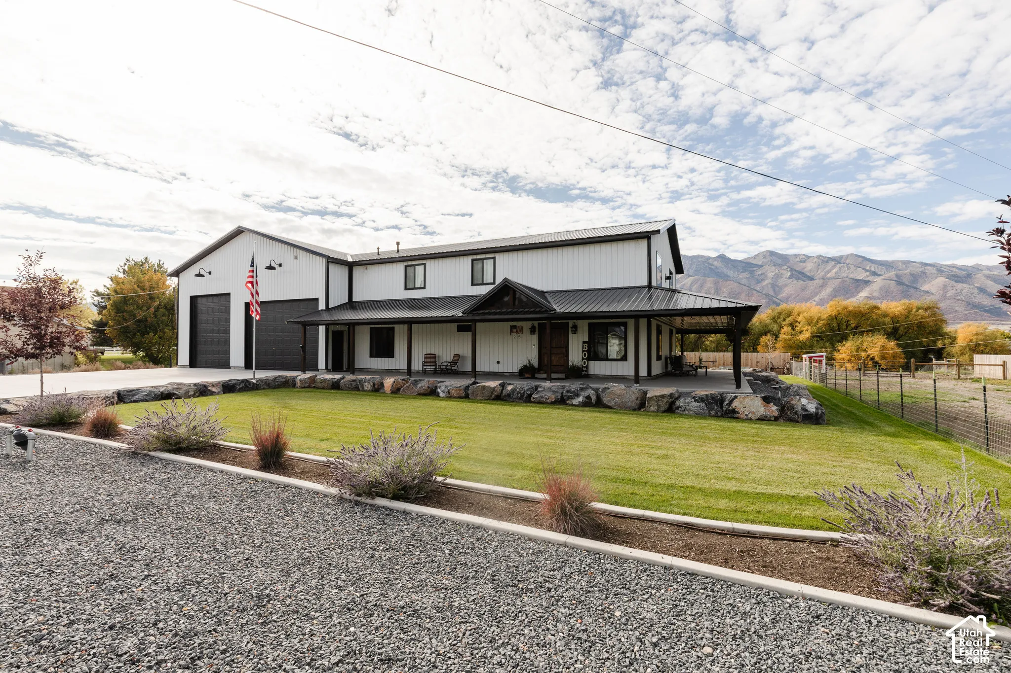 Modern farmhouse exterior featuring vertical metal siding, a covered porch with a metal roof, two-car garage, and manicured landscaping with rock retaining walls against a mountain backdrop