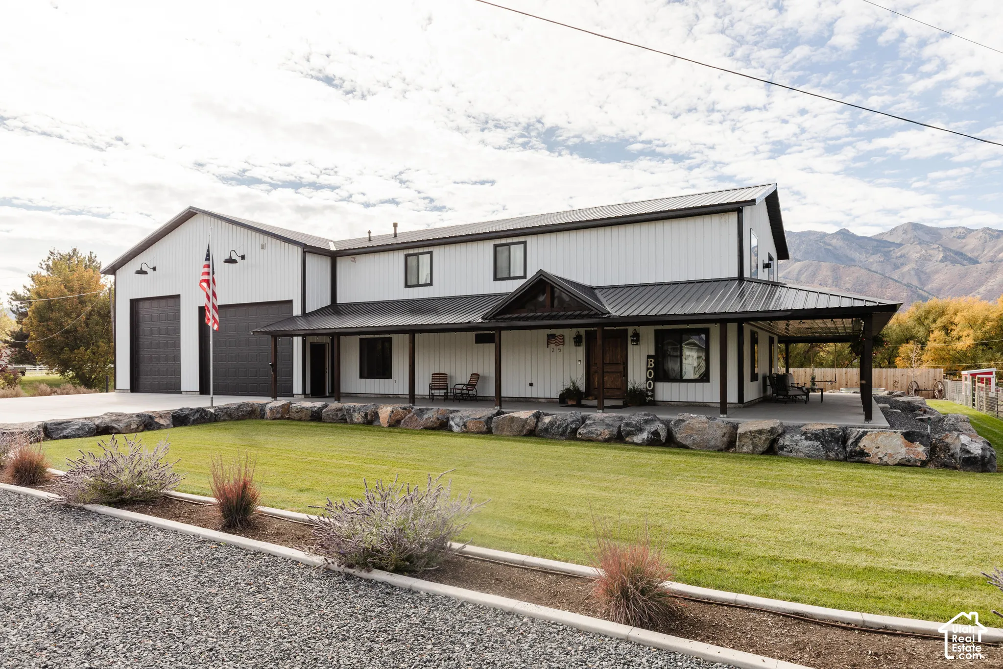 Modern metal-sided exterior featuring a covered wraparound porch, dual-bay garage, stone boulder retaining wall, and mountain backdrop