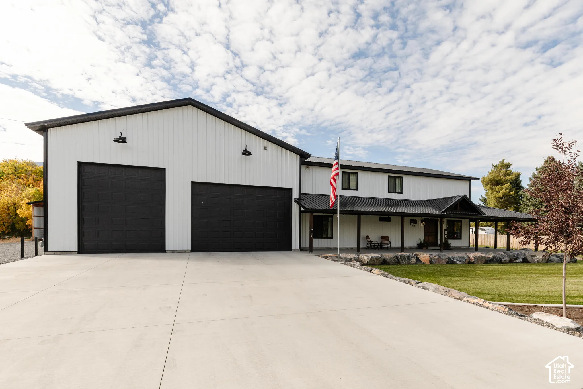 Exterior featuring vertical white metal siding, black garage doors, a covered porch with metal roofing, and a concrete driveway