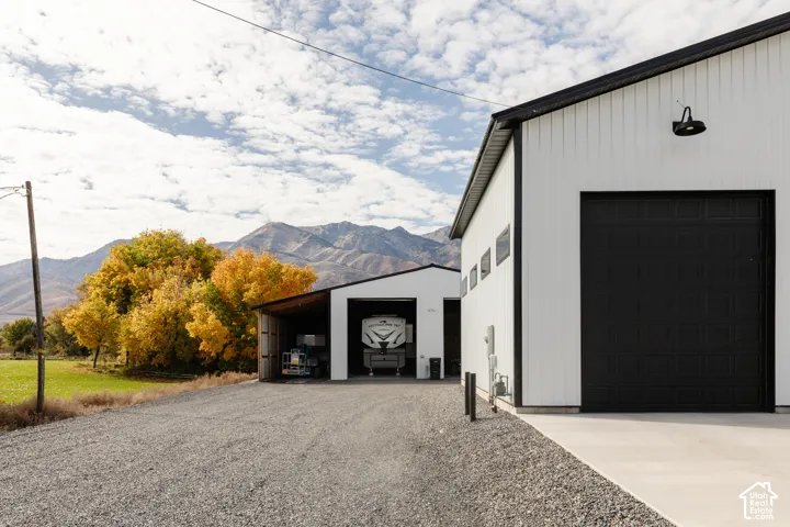Metal-sided detached structures featuring high-clearance overhead garage doors, exterior wall-mounted gooseneck lighting, a gravel driveway, and a mountain backdrop