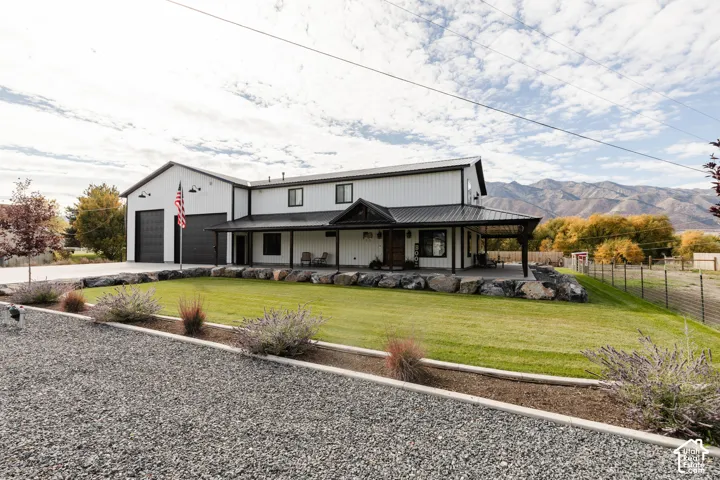 Modern farmhouse exterior featuring vertical metal siding, a covered porch with a metal roof, two-car garage, and manicured landscaping with rock retaining walls against a mountain backdrop
