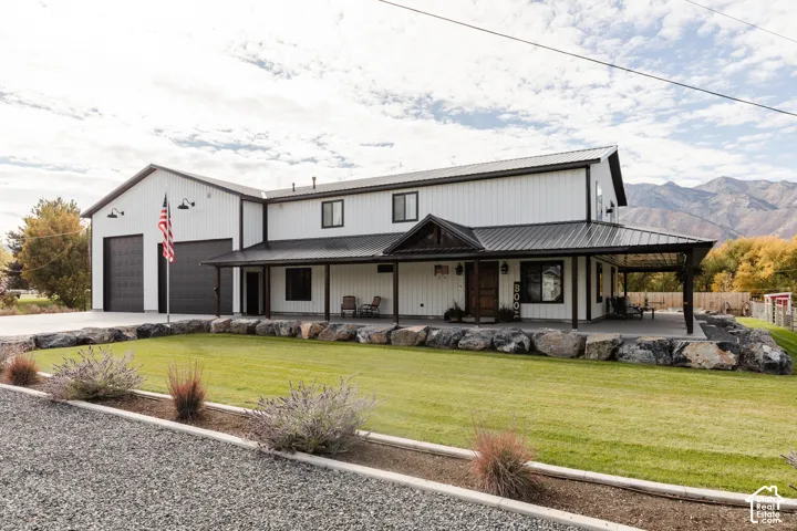 Modern metal-sided exterior featuring a covered wraparound porch, dual-bay garage, stone boulder retaining wall, and mountain backdrop