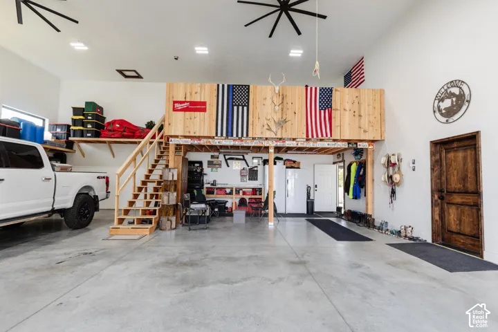 High-ceiling garage space featuring a mezzanine level, built-in wooden staircase, concrete flooring, dual industrial-style ceiling fans, and a solid wood door with decorative hardware