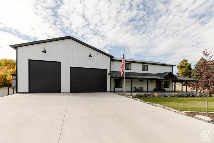 Exterior featuring vertical white metal siding, black garage doors, a covered porch with metal roofing, and a concrete driveway