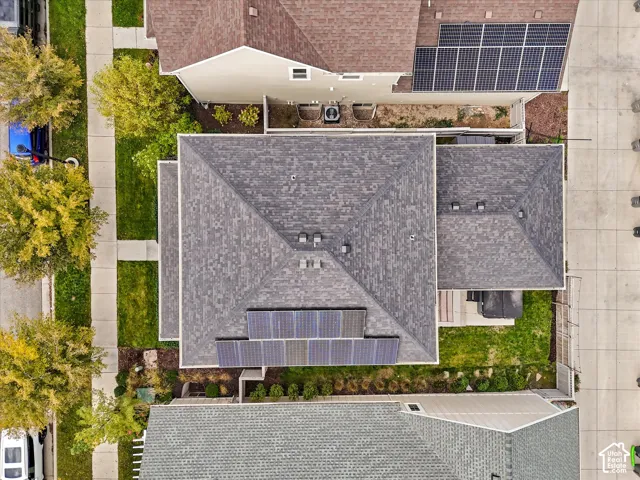 Aerial perspective of the property featuring a hip roof with asphalt shingles, integrated rooftop solar panels, a concrete driveway, and landscaped side and rear lawn areas