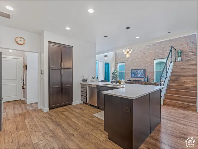 Kitchen featuring dark cabinetry, expansive stone-surface island with integrated stainless steel dishwasher, wood-finish flooring, exposed brick wall, and staircase with wood treads and metal railing