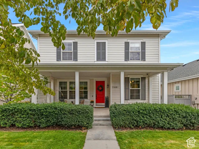 Two-story exterior featuring light-toned horizontal siding, a covered front porch with square columns, a vibrant red entry door, and dark grey decorative shutters