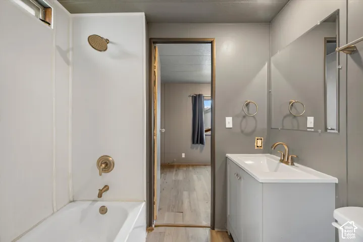 Bathroom featuring a tub-shower combination with brass-finish fixtures, a vanity with a white integrated sink, a wall-mounted mirror, and wood-finish flooring
