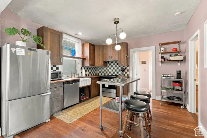 Kitchen featuring wood-finish cabinetry, an apron-front sink, stainless steel appliances, a checkerboard tile backsplash, and wood-finish flooring
