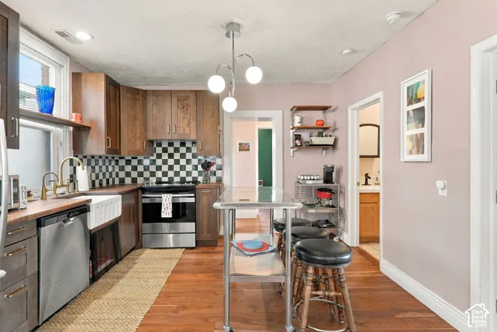 Kitchen featuring wood-finish flooring, apron-front farmhouse sink, stainless steel appliances, checkered tile backsplash, and dark wood cabinetry