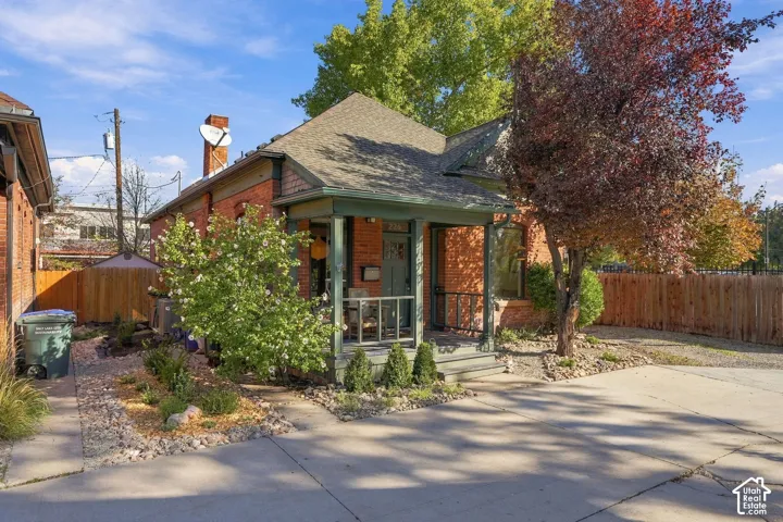 Red brick exterior featuring a covered front porch, shingled roof, mature landscaping, and concrete driveway